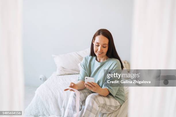 adult brunette woman checking cardiogram on internet using smartphone on bed at home - brunette woman bedroom stock-fotos und bilder