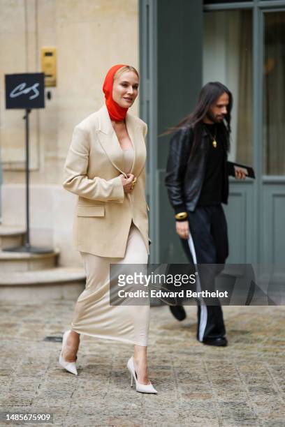 Diane Kruger wears a neon red balaclava, a beige oversized long blazer jacket, a matching beige V-neck long dress, gold rings, a brown shiny leather...