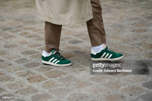 Guest wears white socks, a beige buttoned long trench coat, brown suit pants, green suede and beige logo leather sneakers from Adidas , outside the...