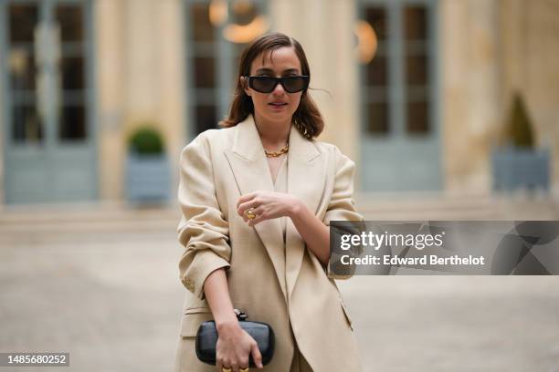 Beatrice Granno wears black sunglasses, a gold large chain necklace, a beige linen V-neck / buttoned suit gilet, a matching beige linen blazer...