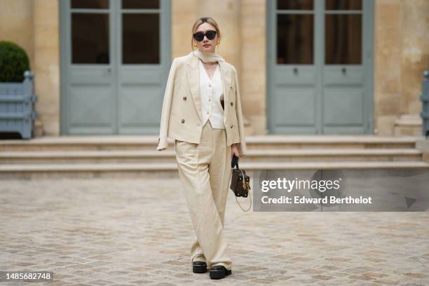 Guest wears black sunglasses, gold earrings, a white silk scarf, a gold chain pendant necklace, a white tweed buttoned V-neck gilet jacket, a beige...