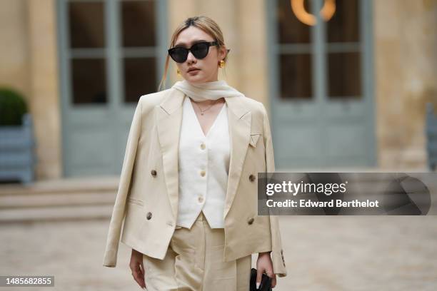 Guest wears black sunglasses, gold earrings, a white silk scarf, a gold chain pendant necklace, a white tweed buttoned V-neck gilet jacket, a beige...