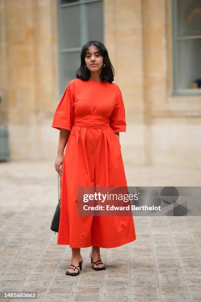 Guest wears silver earrings, a neon red bat short sleeves / large belted / long dress, a black shiny leather puffy quilted handbag, black shiny...