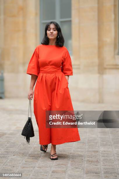 Guest wears silver earrings, a neon red bat short sleeves / large belted / long dress, a black shiny leather puffy quilted handbag, black shiny...