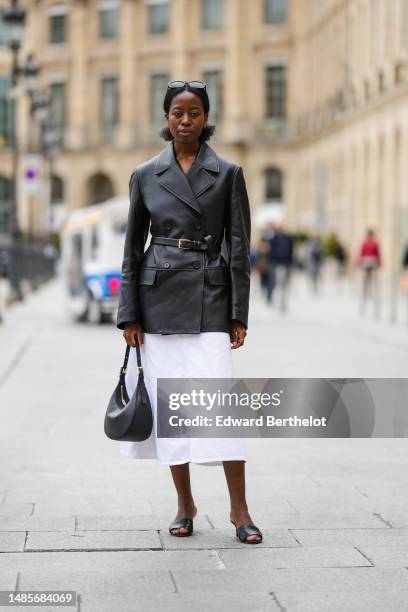 Guest wears black and brown marble print pattern sunglasses, a black shiny leather belted jacket coat, a white midi dress, a black shiny leather...