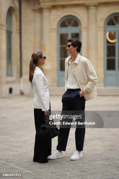 Emelie Lindmark wears black sunglasses, gold earrings, a gold necklace, a white latte buttoned blazer jacket, black suit pants, a black fabric...