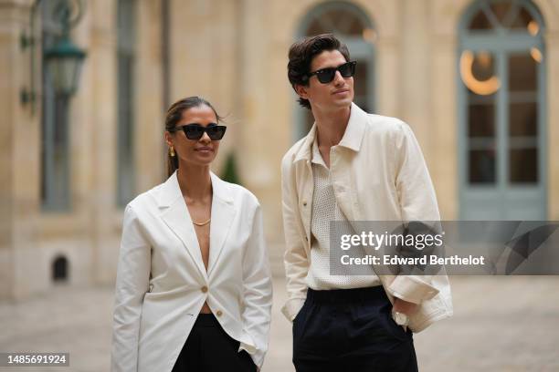 Emelie Lindmark wears black sunglasses, gold earrings, a gold necklace, a white latte buttoned blazer jacket, black suit pants, Moritz Hau wears...