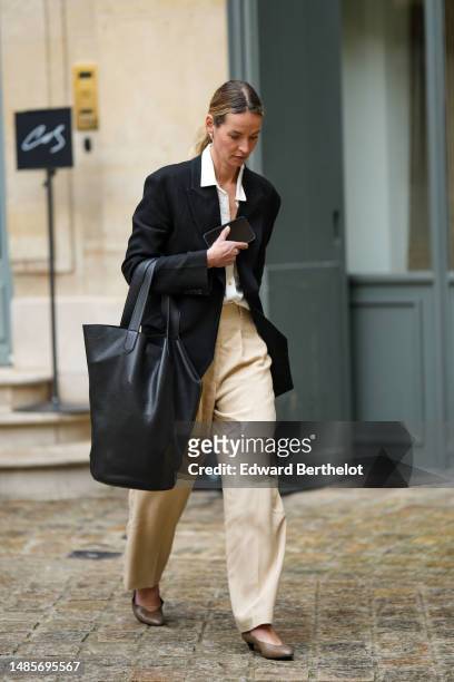 Guest wears silver earrings, a white shirt, a black long blazer jacket, a black shiny leather oversized handbag, beige flared suit pants, brown shiny...