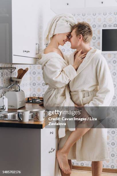 a couple in white terry bathrobes hugging in the kitchen - leg kissing foto e immagini stock