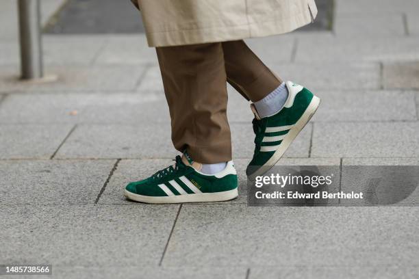 Guest wears a beige buttoned long trench coat, brown suit pants, green suede and beige logo leather sneakers from Adidas , outside the COS show, on...