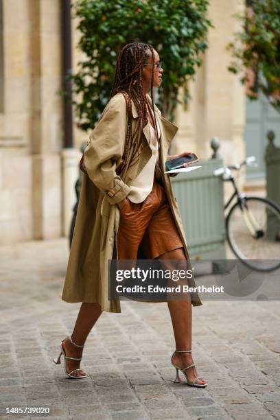 Guest wears black sunglasses, a beige linen buttoned gilet, a pale khaki long trench coat, camel shiny leather suit shorts, gray satin with...
