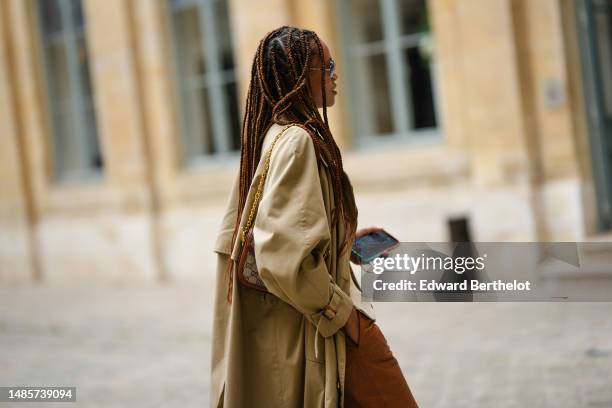 Guest wears black sunglasses, a beige linen buttoned gilet, a pale khaki long trench coat, camel shiny leather suit shorts, outside the COS show, on...