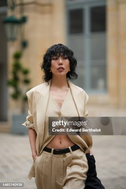 Xiayan Guo wears gold earrings, a beige linen buttoned suit gilet, a beige linen blazer jacket, a matching beige linen suit pants, a black shiny...