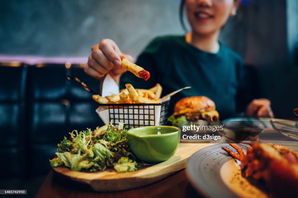 Cropped shot of young Asian woman eating freshly made delicious burger, dipping fries in ketchup on the dining table. Enjoying a feast in the restaurant. People, food and lifestyle