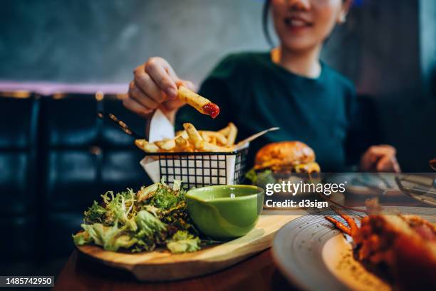 cropped shot of young asian woman eating freshly made delicious burger, dipping fries in ketchup on the dining table. enjoying a feast in the restaurant. people, food and lifestyle - sentarse a comer fotografías e imágenes de stock