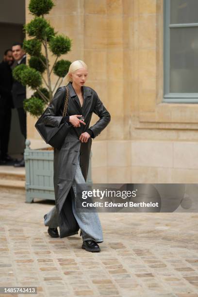 Guest wears a black shoulder-off top, a black shiny leather belted long coat, blue faded denim large pants, a black shiny leather with gold large...