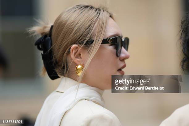 Guest wears black sunglasses from Chanel, gold earrings, a white silk scarf, a gold chain pendant necklace, a white tweed buttoned V-neck gilet...