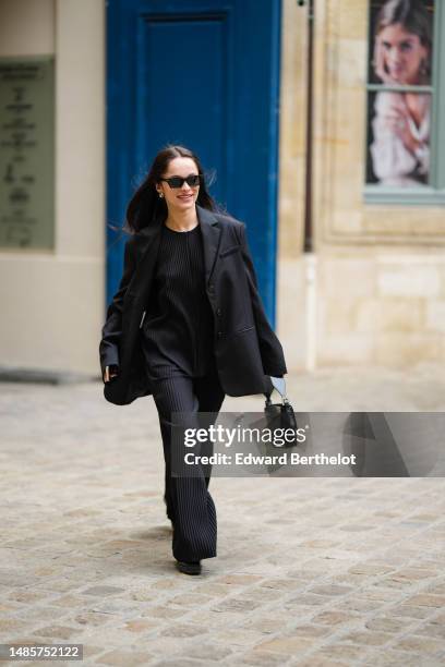 Guest wears black sunglasses, silver earrings, a black pleated / accordion zipper oversized blouse, a black blazer jacket, black suit pants, a black...