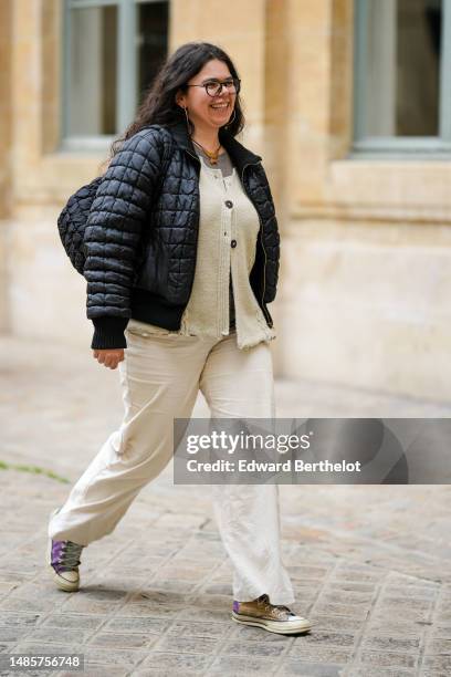 Guest wears black glasses, a gray t-shirt, a beige buttoned wool cardigan, a black quilted pattern bomber coat, a black ruffled fabric shoulder bag,...