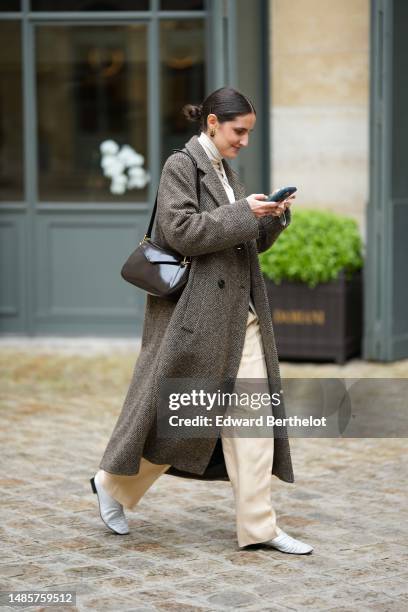 Guest wears a black large hair clip, gold earrings, a white latte turtleneck pullover, a white blazer jacket, a brown wool long oversized coat, pale...