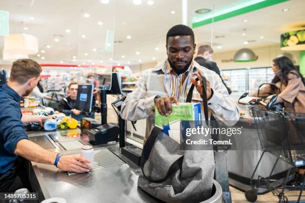 man putting groceries in bag while at supermarket checkout - checkout conveyor belt stock pictures, royalty-free photos & images