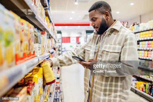 mann, der beim auswählen von artikeln im supermarkt auf das smartphone schaut - supermarkt stock-fotos und bilder