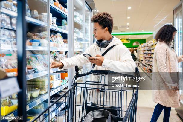 young man looking in fridge while shopping for groceries with mother - supermarket freezer stock pictures, royalty-free photos & images