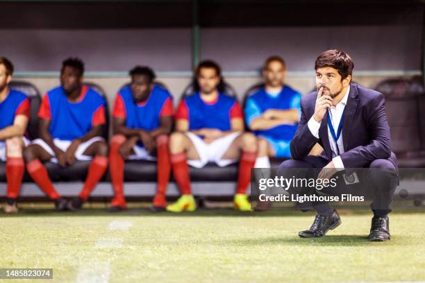 male soccer coach crouches down on a field in front of his reserve players to focus on watching his team play - soccer bench stock pictures, royalty-free photos & images