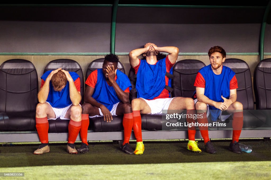 Benched professional soccer players sit with their head in their hands while watching their teammates lose