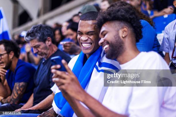 two young adult males laugh at something on a phone while sitting in stadium crowd waiting for soccer game to begin - stadium football stock pictures, royalty-free photos & images