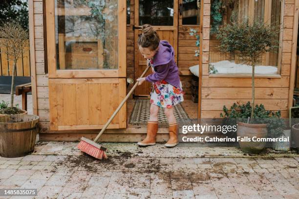 a child uses a yard brush to sweep soil off a paved area outside a greenhouse - organic gardening stock pictures, royalty-free photos & images