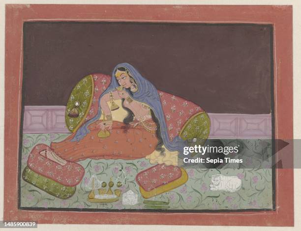 On a green terrace with pink flowers and a pink fence, a lady sits against a large roll pillow, sipping from a glass. On her lap an almost empty...