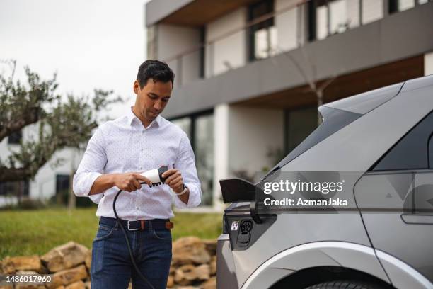 ein gutaussehender erwachsener gemischter mann, der sich darauf vorbereitet, sein elektroauto vor einem modernen gebäude aufzuladen - batteriespeicher stock-fotos und bilder