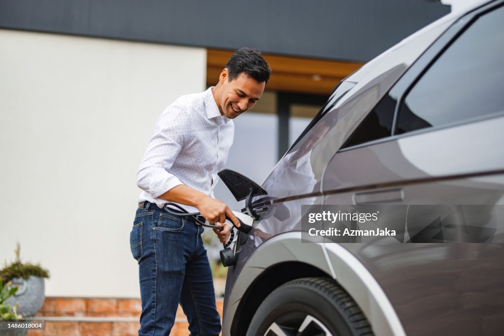 A Happy Handsome Adult Male Charging His Expensive Electric Car Before Leaving His House For A Date