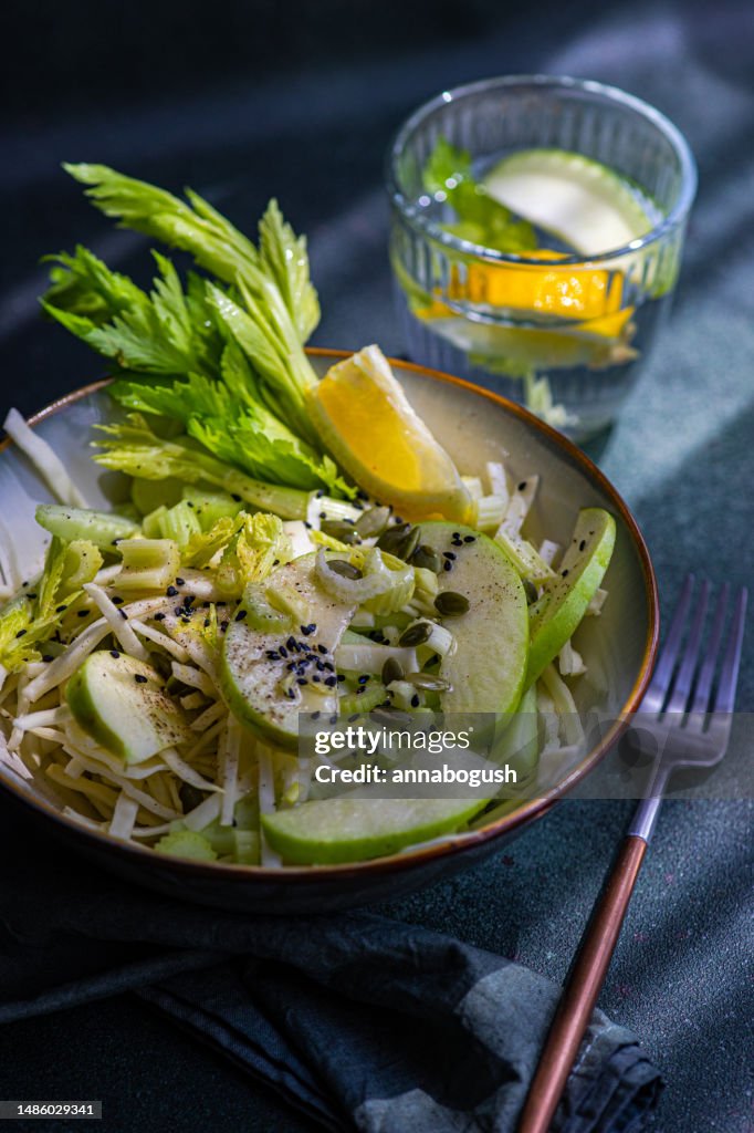 Bowl of celery salad with apples and sesame seeds with a glass of water with apple, celery and lemon