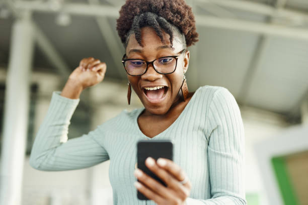 cheerful black woman reading a surprising text message on mobile phone. - black woman phone shock stock pictures, royalty-free photos & images