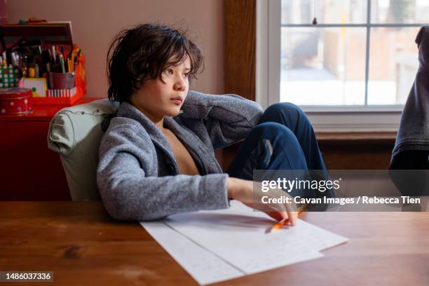 a boy in cardigan sits at table by window working on homework - distraído imagens e fotografias de stock