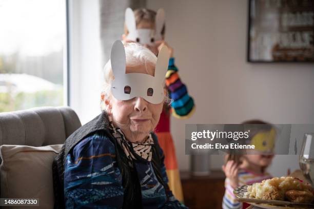 grandmother wearing a novelty easter bunny mask and pulling a pouting facial expression whilst sitting for lunch at a dining table, with two happy grandchildren wearing novelty masks in the background. - easter mask stock-fotos und bilder