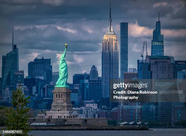 view of the statue of liberty and the empire state building with the late afternoon light shining down. - new-york-city photos et images de collection