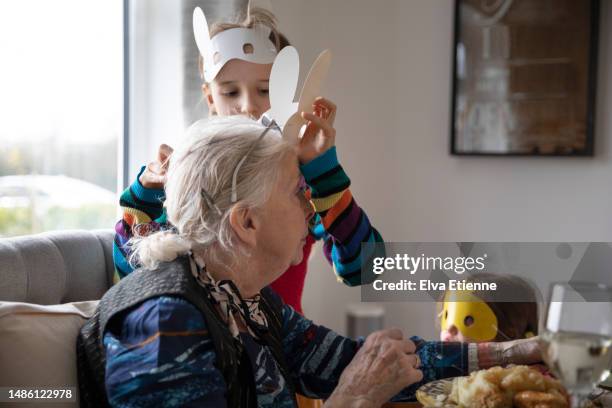 child mischievously putting a novelty easter rabbit mask onto her grandmother during a meal at a dining table, watched by a younger child wearing a yellow chick face mask. - easter mask stock pictures, royalty-free photos & images