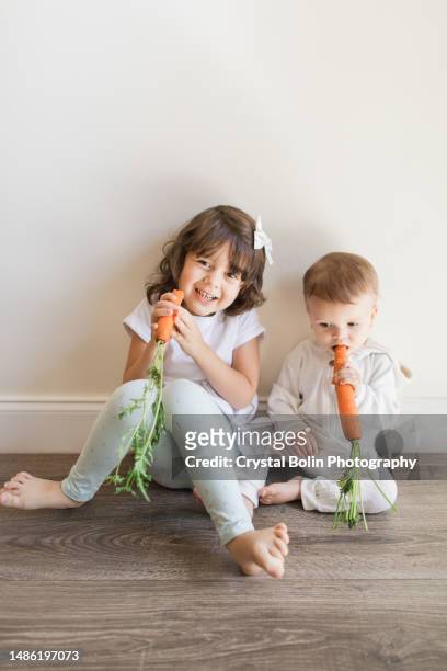 a 4-year-old cuban-american girl with brown curly hair eating a whole carrot with her 15-month-old  baby brother wearing a white bunny outfit, barefoot while at home in a neutral simple space in the spring of 2023 - costume beige stock pictures, royalty-free photos & images