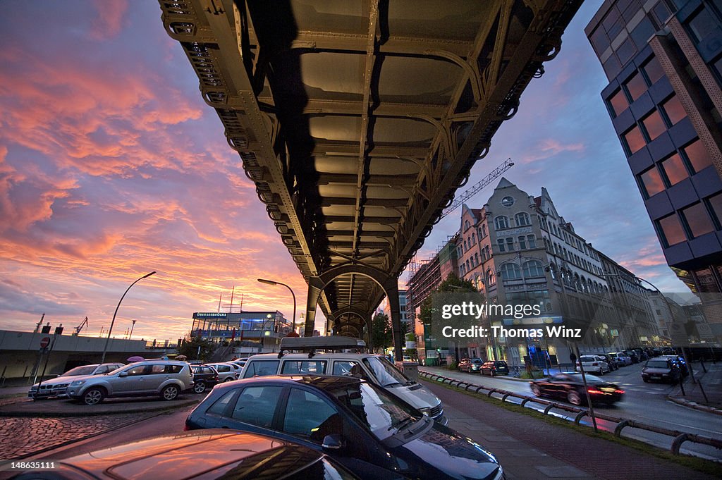 Cars parked under train overpass near Landungsbruecken at dusk.