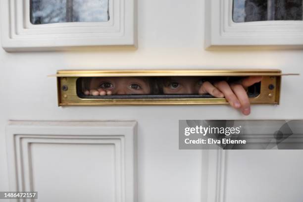 two children peeking out through the narrow space of a gold coloured letterbox on a white painted front door. - brevlåda bildbanksfoton och bilder