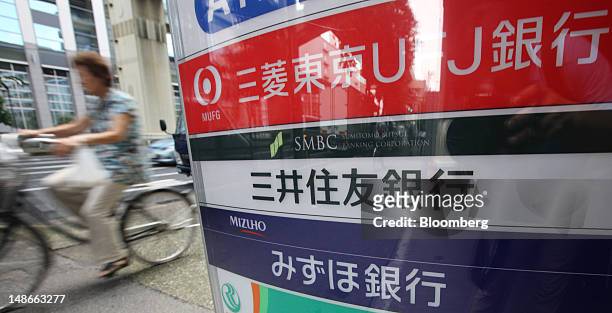 Woman cycles past signs for Bank of Tokyo Mitsubishi UFJ, top, Sumitomo Mitsui Banking Corp., center, and Mizuho Bank Ltd. Outside a convenience...
