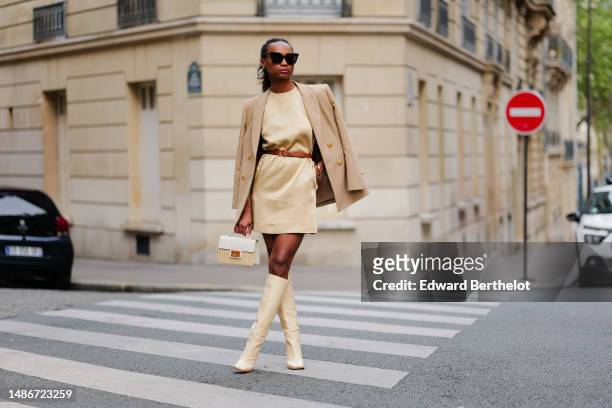 Emilie Joseph wears black large sunglasses, a pale yellow high neck / sleeveless / short dress, a camel shiny leather small belt, a beige with gold...