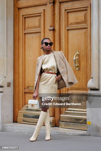 Emilie Joseph wears black large sunglasses, a pale yellow high neck / sleeveless / short dress, a camel shiny leather small belt, a beige with gold...