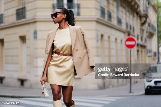 Emilie Joseph wears black large sunglasses, a pale yellow high neck / sleeveless / short dress, a camel shiny leather small belt, a beige with gold...