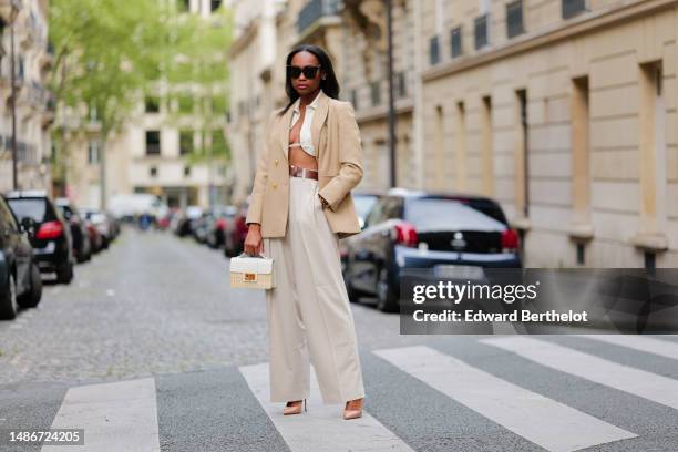 Emilie Joseph wears black large sunglasses, small earrings, a white linen V-neck / cropped laces shirt, a beige with gold buttons blazer jacket, a...