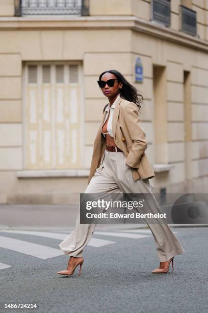 Emilie Joseph wears black large sunglasses, small earrings, a white linen V-neck / cropped laces shirt, a beige with gold buttons blazer jacket, a...