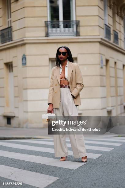 Emilie Joseph wears black large sunglasses, small earrings, a white linen V-neck / cropped laces shirt, a beige with gold buttons blazer jacket, a...